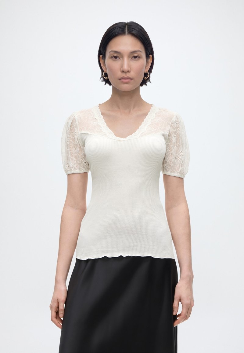 Femme aux cheveux noirs courts portant un haut blanc côtelé avec des manches bouffantes en dentelle et une jupe noire, debout devant un fond blanc uni.