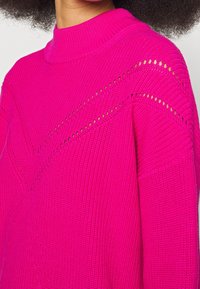 Maglione fucsia a maglia con colletto alto, caratterizzato da motivi a zigzag testurizzati e dettagli di pizzo sulle spalle.