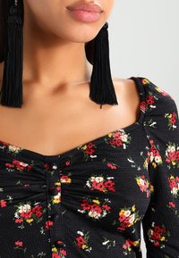 Svart blommig topp med röda, gula och vita blommor, med texturerad väv, en sweetheart-uteskärning och dekorativa knappdetaljer. Svarta fransörhängen.