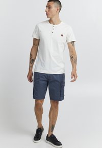 Kortermet hvit bomulls-T-skjorte med tre knapper, kombinert med marineblå cargo-shorts og svarte sneakers. Tatoveringer synlige på armene.