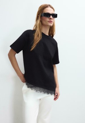 Femme portant un haut noir à manches courtes avec bordure en dentelle, pantalon blanc et lunettes de soleil rectangulaires noires, debout devant un fond blanc.