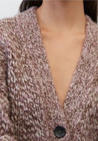 Cardigan vyrobený z tlusté, texturované příze v hnědých a světle růžových tónech s hlubokým výstřihem do V a jedním černým knoflíkem na zapínání.