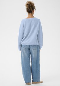 Femme aux cheveux bouclés portant un pull bleu clair, un jean large délavé et des chaussures plates beiges, se tenant de dos sur fond blanc.