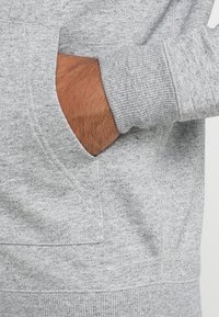 Šedá mikina s kapucí, vybavená klokáním kapsou, žebrovanými manžetami a měkkou, texturovanou tkaninou. Design je jednoduchý bez dalších vzorů nebo zdobení.