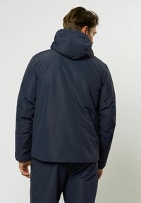 Mann in einer marineblauen wasserdichten Kapuzenjacke und passenden Hosen, von hinten vor einem schlichten hellen Hintergrund dargestellt.