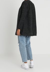 Cappotto oversized in lana nera con maniche lunghe e scollo rotondo, abbinato a jeans chiari strappati e sneaker bianche alte.