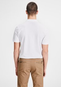 Weißes Kurzarm-Polohemd mit klassischem Kragen, getragen mit hellbraunen Hosen. Der Stoff wirkt glatt und hat eine Standardpassform.