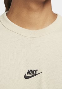 Beige cotton t-shirt with a ribbed crew neck, featuring a black embroidered Nike logo on the chest, smooth texture and relaxed fit.