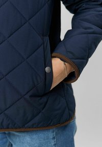 Jūrinė dygsniuota striukė su rudomis detalėmis, turinti užsegamos kišenės dizainą ir tekstūruotą audinį. Artimas kadravimas prie rankogaliai ir rankos kišenėje.