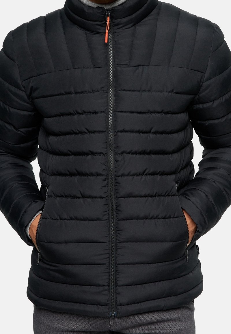 Schwarze gesteppte Daunenjacke mit hohem Kragen, Frontreißverschluss und Seitentaschen. Hat eine glatte Textur und einen kontrastierenden orangefarbenen Reißverschlussgriff.