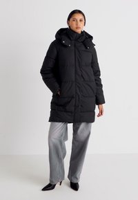 Abrigo acolchado negro con cuello alto y capucha, con diseño acolchado horizontal, cierre de cremallera y bolsillos laterales; llevado sobre pantalones de pierna ancha grises.