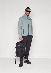Casaco de fleece cinza com bolsos com fecho, calças pretas e uma mochila preta. Possui óculos de sol e sapatos esportivos escuros.