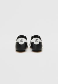 Chaussures en cuir noir avec un talon blanc et un logo, présentant un design incurvé, un système de laçage à l'avant et une semelle en caoutchouc gomme pour l'adhérence.