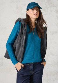 Langärmliges, blau gestreiftes Oberteil unter einer grauen, mit Reißverschluss versehenen Fleeceweste, kombiniert mit dunklen Jeans und einer türkisfarbenen Mütze mit strukturiertem Finish.