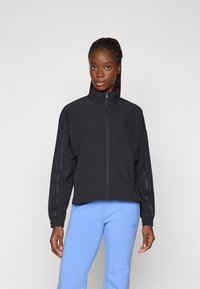 Black zip-up jacket with high collar and ribbed cuffs, featuring three vertical stripes on each sleeve. Worn over light blue pants.