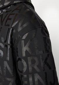 Black hoodie featuring a textured pattern of uppercase letters in varying gloss finishes, with a smooth fabric texture and fitted silhouette.
