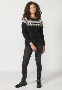 Un pull noir duveteux avec un empiècement clair à motifs. Associé à un jean noir ajusté et des bottes lacées noires. Fond lisse.