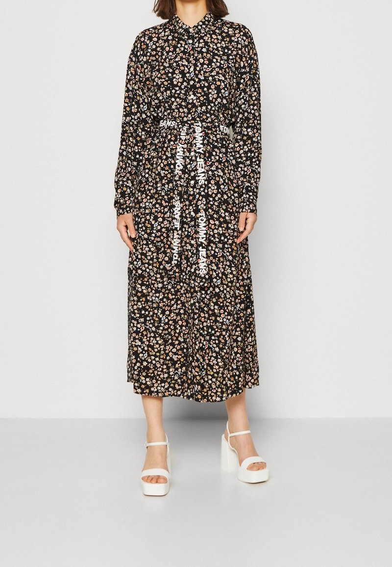 Femme portant une robe midi noire fleurie à manches longues avec une ceinture blanche "Tommy Jeans" et des sandales blanches à plateforme, debout devant un fond uni.
