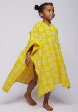 Pige går barfodet iført en gul hættede håndklædeponcho med hvide cirkulære frugtmønstre på en ensfarvet baggrund.