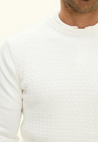Biały sweter z teksturowanym wzorem z przodu, z okrągłym dekoltem i dzianinowym materiałem. Gładkie rękawy i detal przy kołnierzu widoczne.