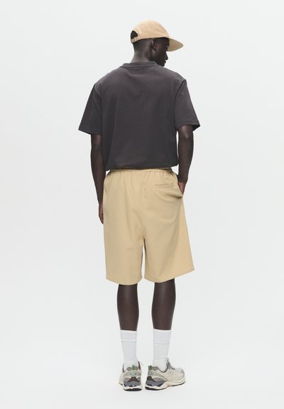 Tenue décontractée composée d'un t-shirt en coton noir, de shorts beiges avec une taille élastique et de baskets gris clair. La casquette est beige.