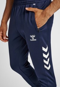 Námornícky modré športové nohavice vyrobené z hladkej tkaniny. Majú biele logo a chevronový vzor na ľavej nohe. Obsahujú bočnú vrecko na zips.