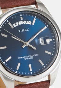 Orologio Timex con quadrante blu, indicatori delle ore in tonalità argento e display giorno/data; cassa in acciaio inossidabile e cinturino in pelle marrone. Impermeabile fino a 5 ATM.