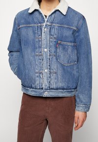 Blå jeansjacka med fuskpälskrage, framskjutande knappar, bröstficka och lätt slitning. Bärs över ett svart plagg och bruna byxor.