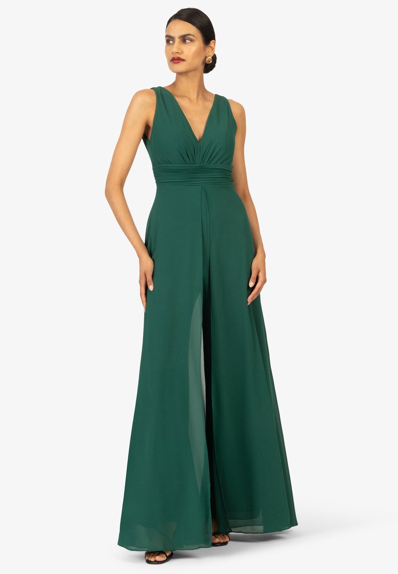 Grüner ärmelloser Jumpsuit mit tiefem V-Ausschnitt, gefaltetem Oberteil, weitem Bein und fließendem Stoff; kombiniert mit schwarzen High Heels und goldenen Ohrringen.