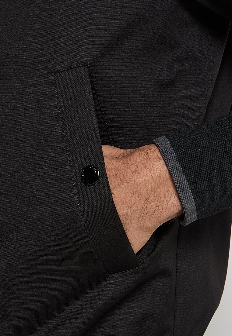 Männliche Hand teilweise in der schwarzen Jackentasche mit einem schwarzen Druckknopf, der Ärmelsaum eines dunklen Ärmels ist vor einem neutralen Hintergrund sichtbar.