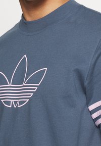 Modrá bavlněná tričko s velkým růžovým trefoil logem a třemi horizontálními růžovými pruhy na rukávech. Kulatý výstřih a měkká textura.