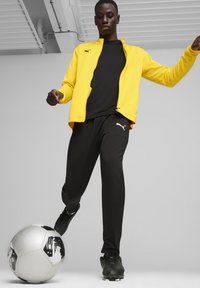Gelbe Zip-Jacke, schwarzes Oberteil und schwarze Hose, kickt einen schwarz-weißen Fußball, elegantes sportliches Design, leichtes Material, sportliche Passform.