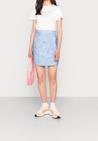 Nainen, jolla on päällään valkoinen graafinen t-paita, sininen marmoroitu mini-hame, valkoiset sukat ja oranssi-valkoiset lenkkarit, kantaa pinkkiä käsilaukkuja ja seisoo valkoista seinää vasten.