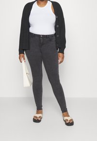 Cárdigan negro acanalado, camiseta blanca ajustada, jeans skinny gris oscuro, sandalias beige con tiras, sosteniendo un pequeño bolso blanco.