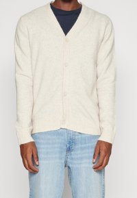 Krämfärgad kofta i mjukt material, med V-ringning, ribbad nederkant och ärmslut, samt fem knappar framtill.