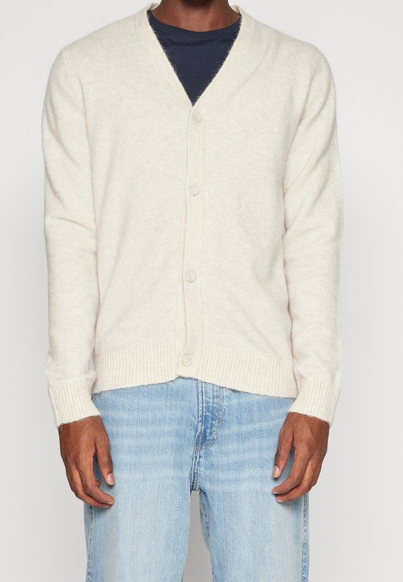 Krämfärgad kofta i mjukt material, med V-ringning, ribbad nederkant och ärmslut, samt fem knappar framtill.