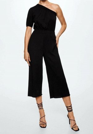 Kvinde iført en sort jumpsuit med én skulder, brede cropped ben og sorte sandaler med høje hæle og stropper mod en ensfarvet baggrund.
