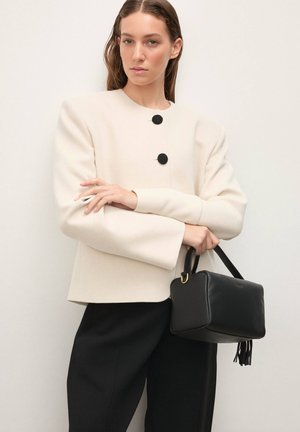 Kreminės spalvos ilgarankovė striukė su juodais mygtukų detalėmis, derinama su juoda odine rankine su dirželiu ir kutais. Juodos kelnės.