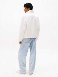 Witte, fluffy fleece jack met een hoge kraag, gecombineerd met lichtblauwe, loszittende jeans en witte sneakers. Effen achtergrond.