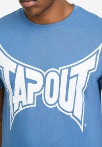Hellblaues Baumwoll-T-Shirt mit großem weißem "TAPOUT"-Logo auf der Brust, mit Rundhalsausschnitt und kurzen Ärmeln.