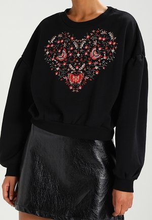 Sweat-shirt noir à manches longues avec broderie rouge florale et papillon formant un cœur, associé à une mini-jupe noire brillante.