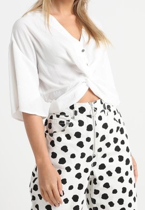 Žena nosící bílý krátký top na knoflíky a bílé kalhoty s vysokým pasem, zdobené velkými nepravidelnými černými puntíky a předním zipem.