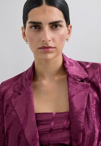 Una mujer con cabello oscuro y liso, y pendientes de aro dorados, lleva un blazer texturizado morado y un top de tubo a juego, contra un fondo claro y liso.