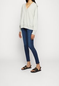 Sudadera de cuello en V gris claro, de corte relajado, con mangas largas; combinada con jeans ajustados azul oscuro y sandalias negras de tiras.