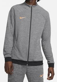 Šedá zipová bunda s texturovanou látkou, černé akcenty na manžetách a lemu, se světlou oranžovou Nike logem na levé hrudi.