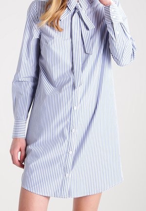 Femme portant une robe-chemise à manches longues rayée verticalement bleue et blanche, avec un nœud noué au niveau du col, boutonnée sur le devant.