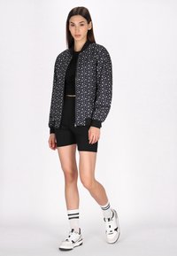 Sort bomberjakke med hvidt stjernesystem, ribbede manchetter og halsudskæring. Sammen med en sort crop top, shorts og hvide sneakers med sorte detaljer.