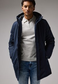 Chaqueta acolchada azul marino con capucha, que presenta un cierre frontal con cremallera y botones a presión, llevada sobre un polo gris claro y jeans azul claro.