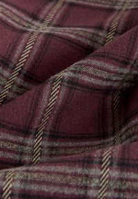 Kariertes Gewebe in Burgunderrot und Grau, hergestellt aus einer weichen Wollmischung. Es verfügt über sich kreuzende Linien in verschiedenen Breiten, die ein strukturiertes Muster erzeugen.