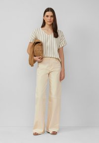 Beige en crème gestreepte top met korte mouwen en een v-hals, gecombineerd met lichtbeige brede broekspijpen. Model houdt een geweven tan tas vast.